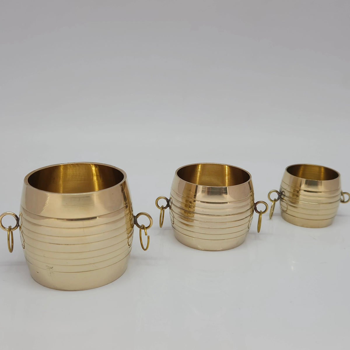 Three small sizes of traditional brass nazhi and para measuring cups lined up side-by-side to showcase the gradual scale variations.

