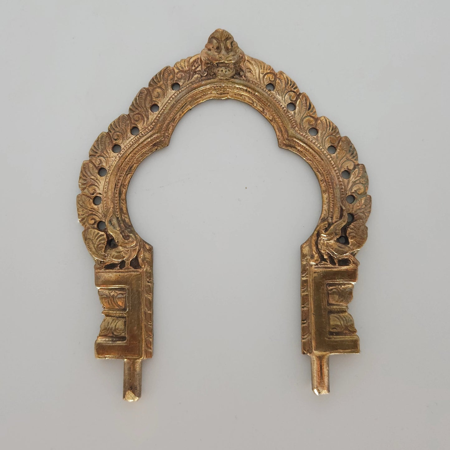 An ornate gold-toned metal arch-shaped decorative piece with intricate cutout detailing and two vertical prongs, shown against a plain light gray background.