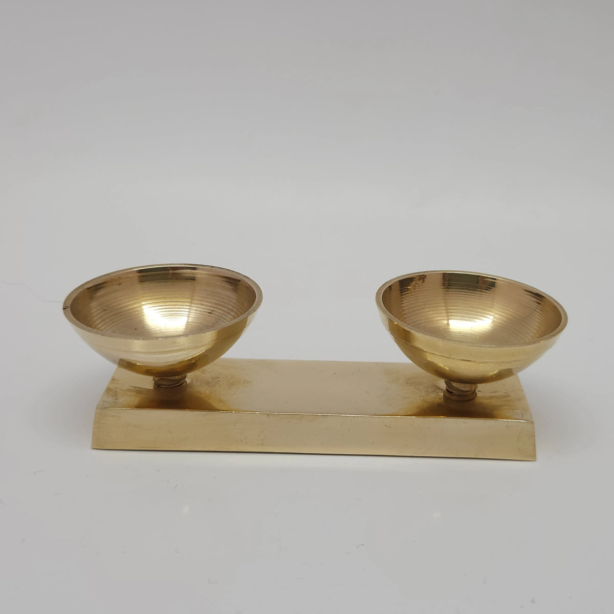 Front profile view of a pure glossy brass twin bowl altar stand focusing on the two connected cups and the thick flat rectangular base plate