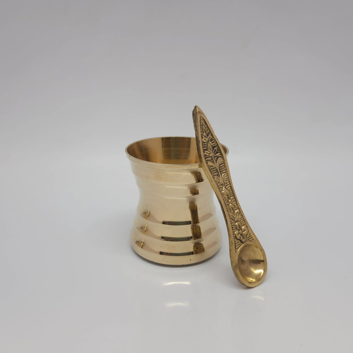 Three-quarter front view of the small size pure brass panchapatra tumbler with its matching leaf-handled udhrani spoon resting outside the rim by Sri Swaminathan and Co