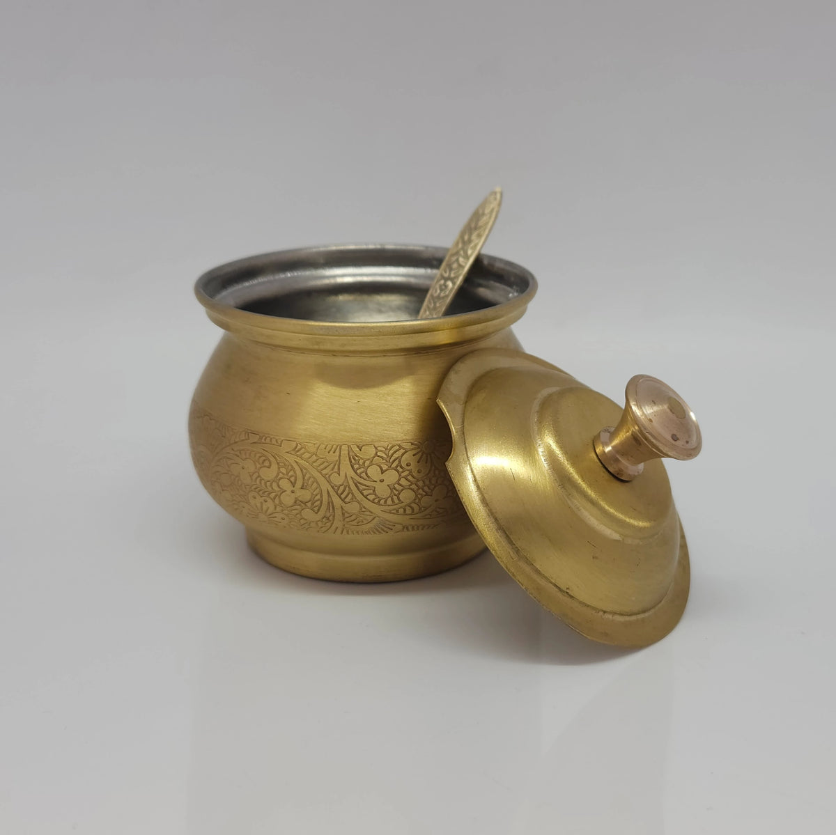 Direct side profile view of the premium brass engraved ghee pot with its lid slightly propped open alongside the accompanying serving spoon