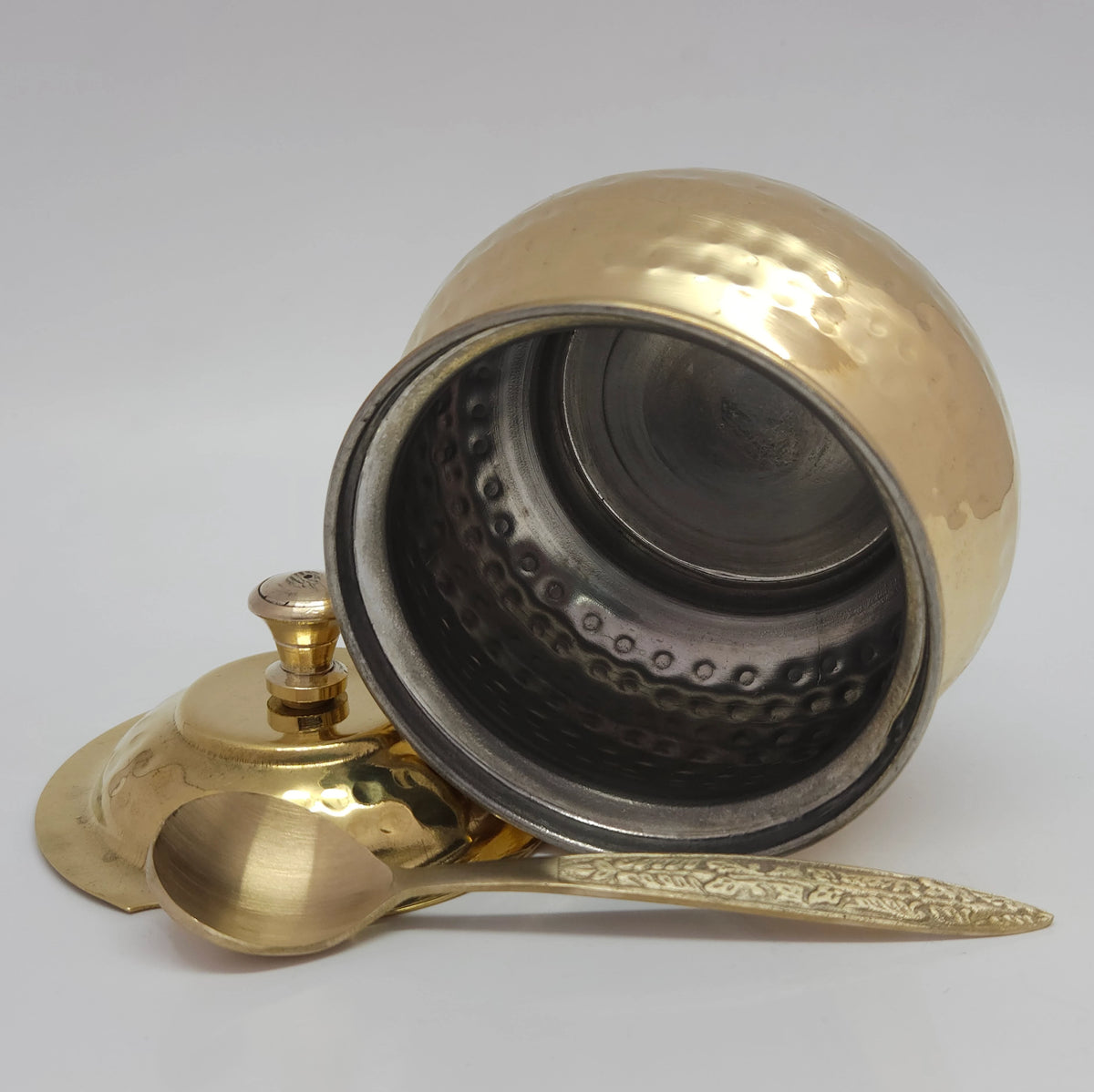 Tilted side perspective view of the glossy hammered brass ghee pot showcasing the contrasting interior silver-colored tin coating safety layer