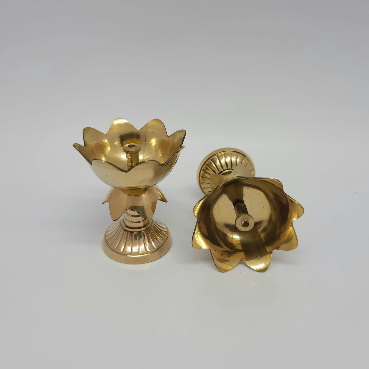 Angled view displaying one brass pedestal lotus tulip diya standing upright next to another diya tilted back to reveal the deep inner oil reservoir capacity.