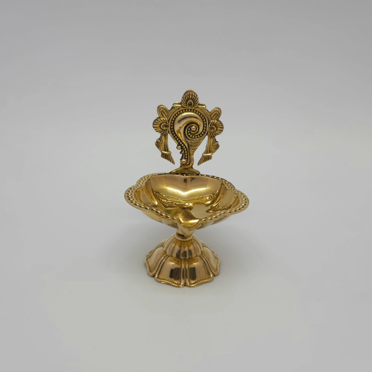 Straight front view of a single solid brass pedestal altar vilakku embossed with the divine Sankhu discus frame.