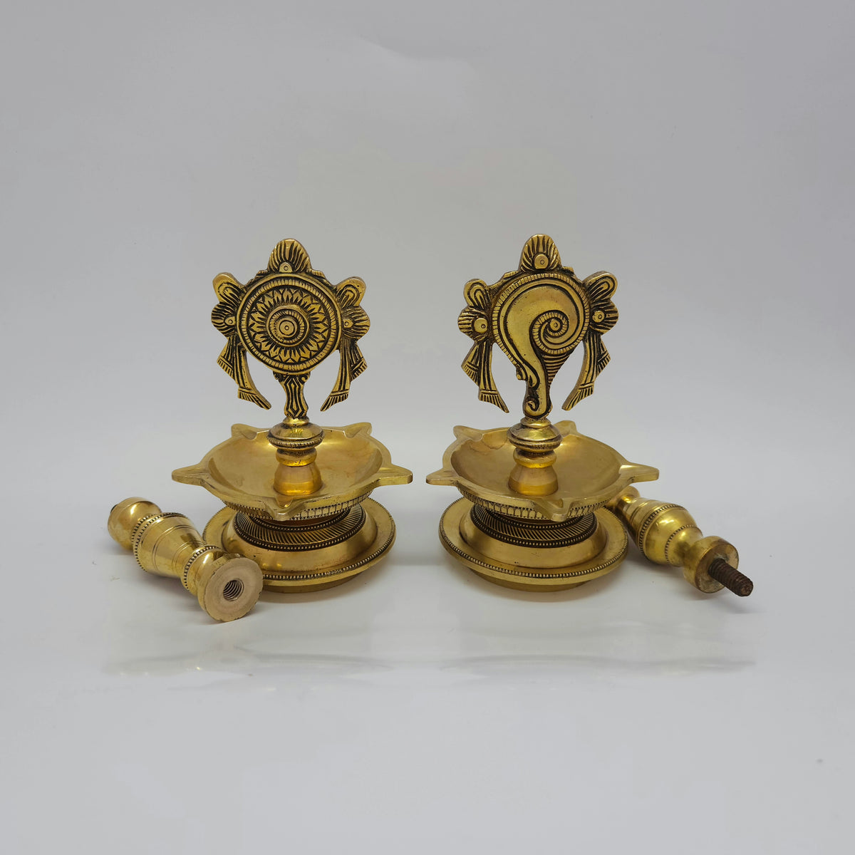 Angled view displaying the brass Shanku and Chakra lamp heads unscrewed from their 25cm standing pedestal stems to showcase the modular threads.