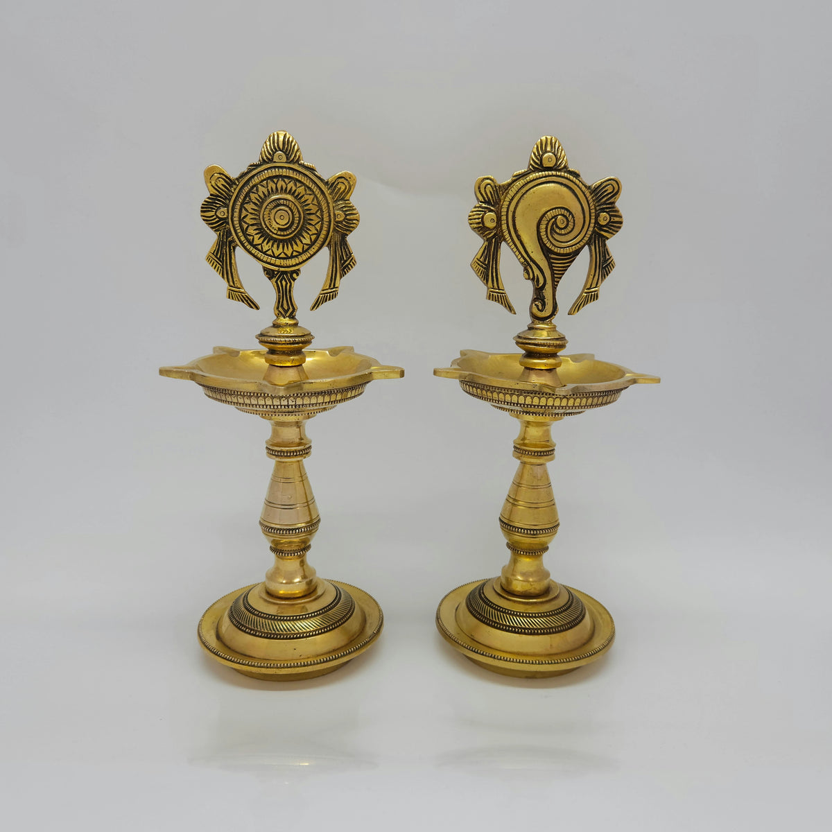Straight front view of a single solid brass 25cm standing kuthu vilakku embossed with the divine Shanku conch backframe by Sri Swaminathan and Co.