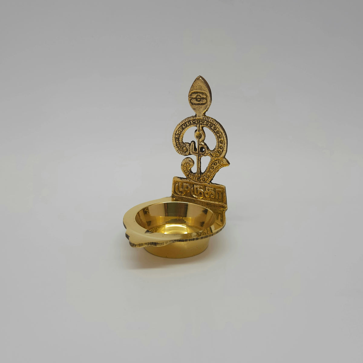 Three-quarter front left perspective view of the 85g solid brass Murugan lamp showing the curved lines of the Tamil Om frame.