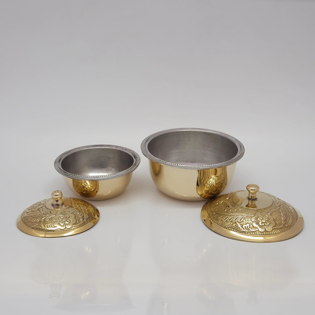 Front view of two sizes of pure brass tin-lined uruli serving bowls with their floral engraved lids placed open on a white surface to show the inner metallic silver lining.
