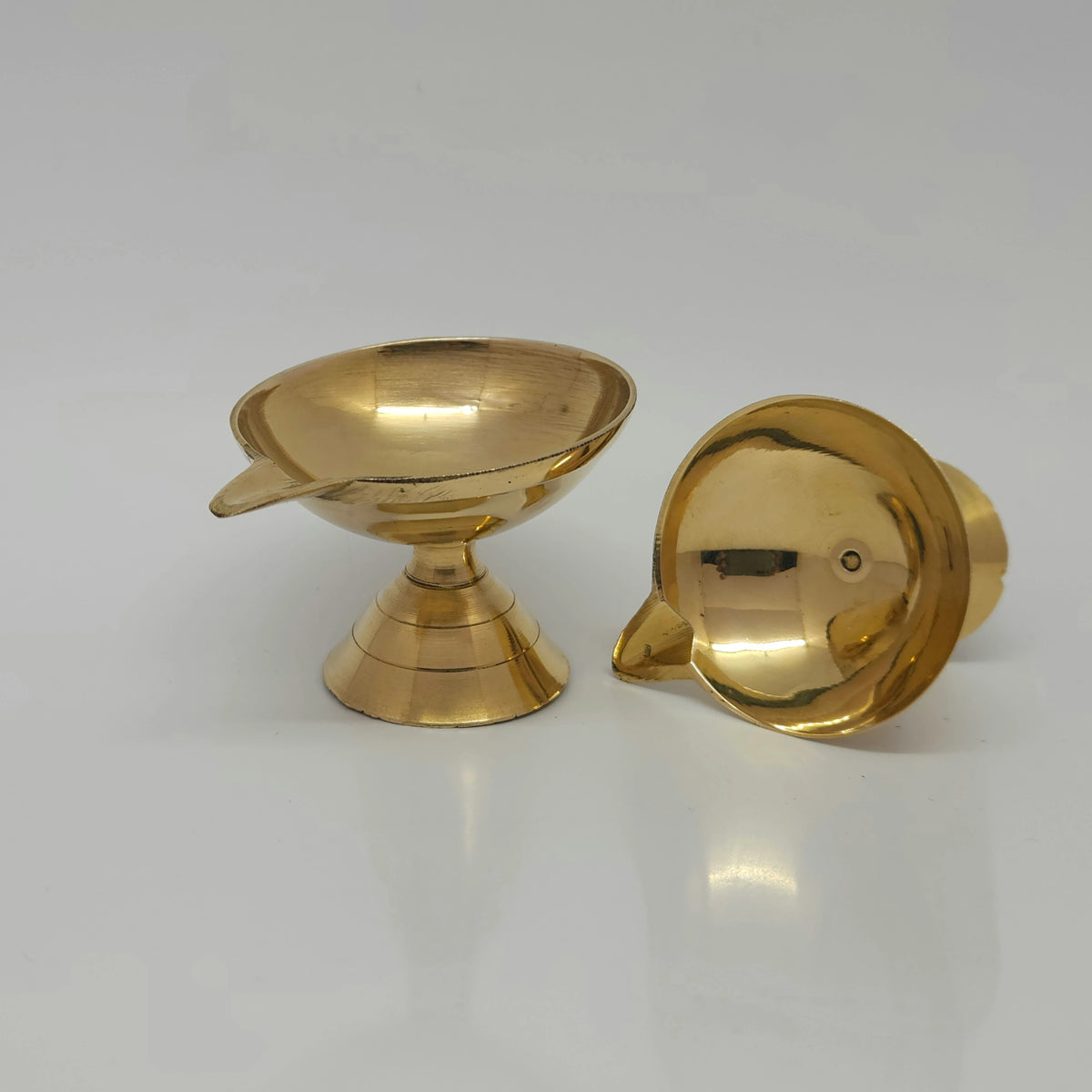 Angled view displaying one assembled brass pedestal diya standing upright next to another diya tilted forward to reveal the inner single-wick oil reservoir depth.