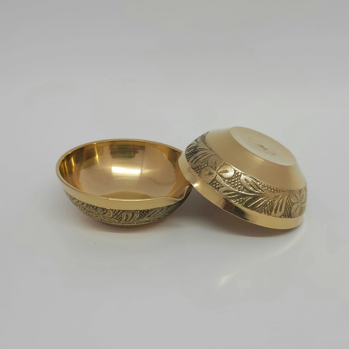 Angled view displaying one floral engraved brass agal vilakku standing upright next to another diya tilted back to reveal the clean 3.4cm flat-bottom layout.