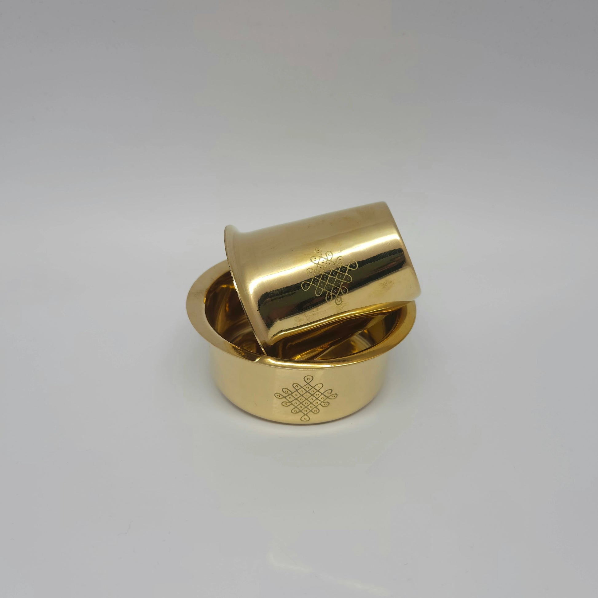 Angled view of the personalized glossy finish brass coffee cup resting sideways on the rim of the traditional 4-Kolam engraved davara basin.