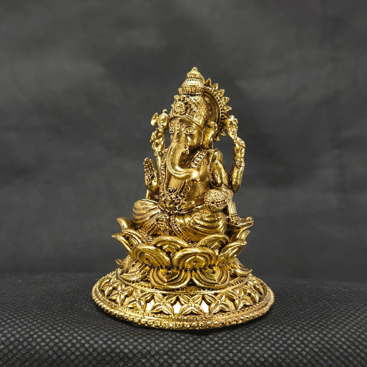 Three-quarter front left perspective view of the two and a half inch brass Ganesha statue on a round lotus peedam highlighting the sharp chiseled lines of his traditional weapon attributes.