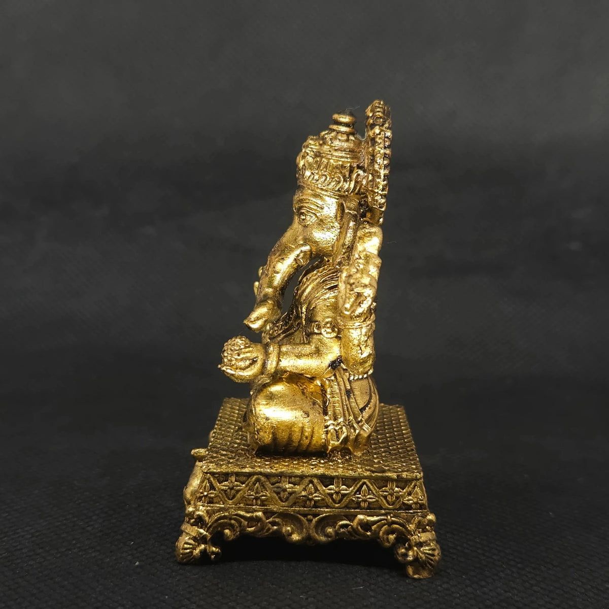 Exact left side profile view of the solid brass Ganesha statue on an ornate peedam showcasing the sharp three-dimensional lines of his weapon attributes and base thickness