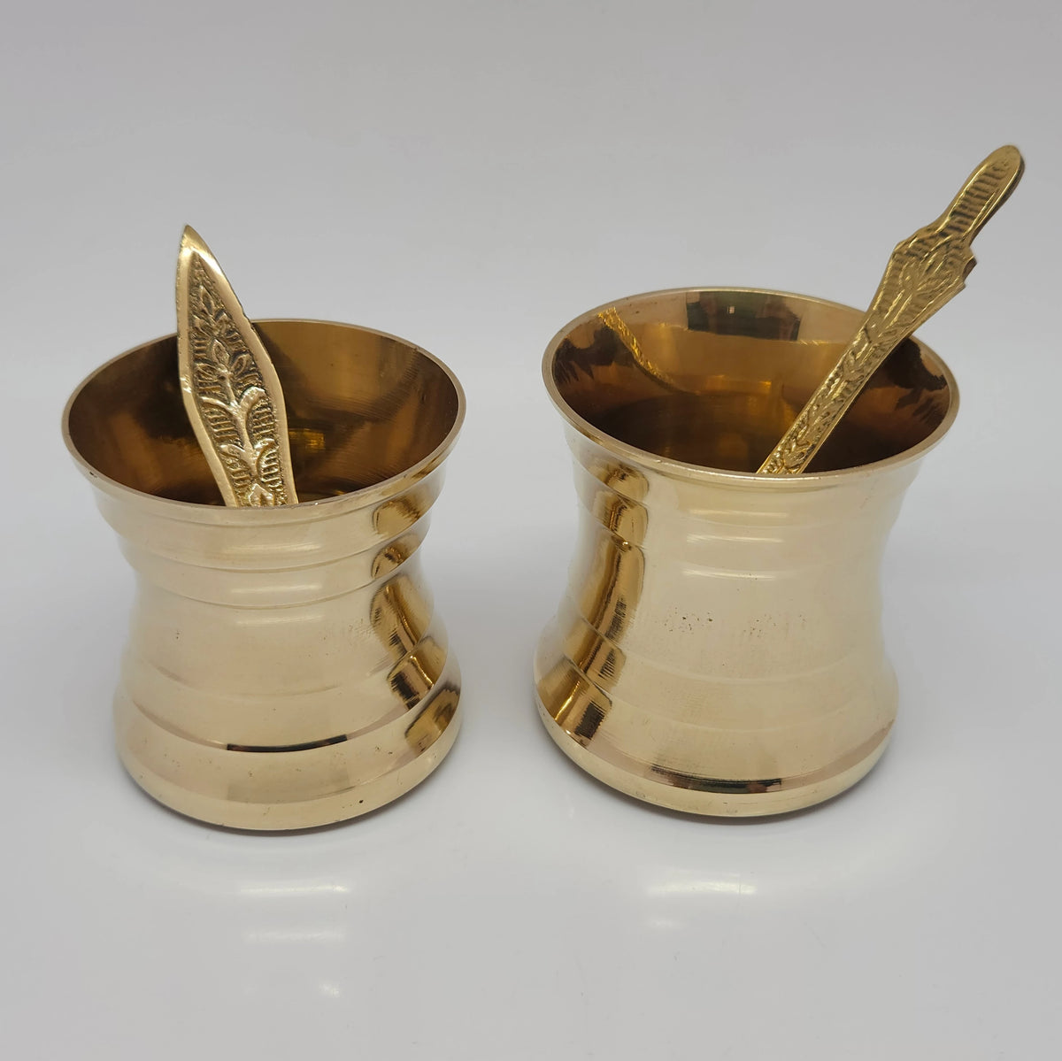 Top-down perspective view of two pure brass theertham cups with spoons showcasing the wide rim diameters and polished interior basins for holding holy water