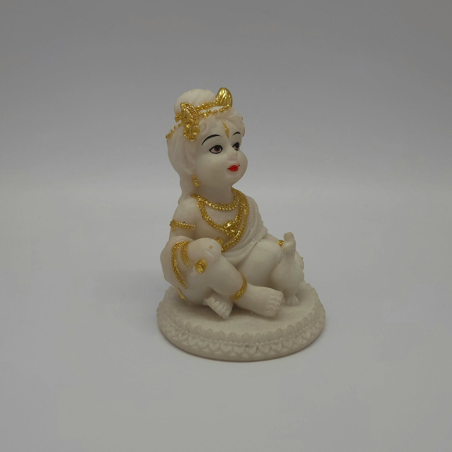 Angled front right look of the white poly-marble child Krishna figurine showcasing his cross-legged sitting posture and gold ornaments.